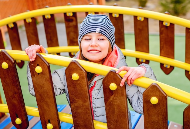 Kleines Mädchen, das hinter Holzzaun mit gelbem Metallhandlauf auf Spielplatz sitzt und lächelt