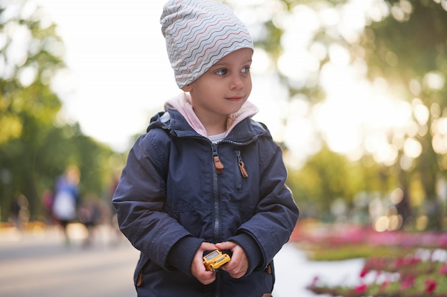 Kleines Mädchen, das Hut warme Herbstjacke trägt, geht durch Herbstpark, der Spielzeugauto hält