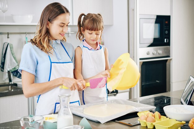 Kleines Mädchen, das ihre Mutter betrachtet, die Kuchenteig auf Backblech gießt