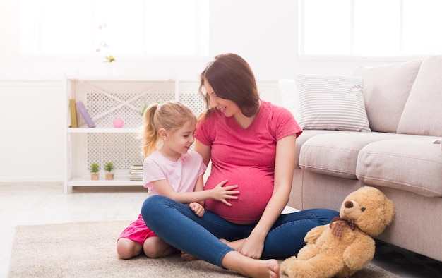 Kleines Mädchen, das ihren schwangeren Mutterbauch berührt Erwartete Mutter, die mit ihrer Tochter zu Hause auf dem Boden sitzt, Spaß hat und Babydrücke fühlt, Platz kopieren