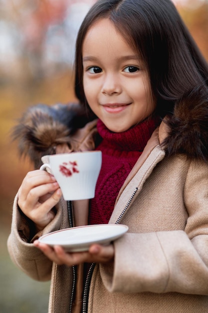 Kleines Mädchen, das im Herbstpark warmen Tee trinkt