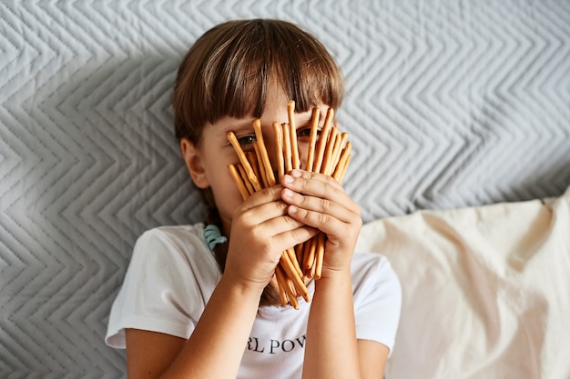 Kleines Mädchen, das in den Händen hält und ihr Gesicht mit Keksstäbchen bedeckt, dunkelhaariges unbekanntes weibliches Kind, das ein weißes, lässiges T-Shirt trägt, auf dem Sofa sitzt und durch Brezeln schaut.