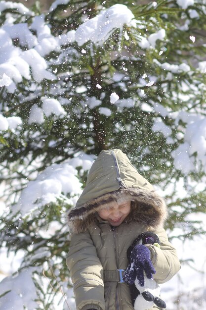 Kleines Mädchen, das in den schneebedeckten Winterwald geht. Kinderfreude im Schnee, der von den Zweigen einer Fichte fällt.