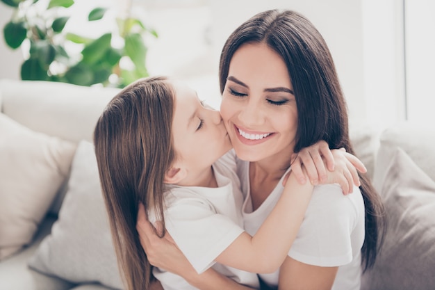 Kleines Mädchen, das junge Mama der Wange küsst, die sie im Haus umarmt