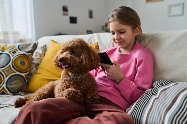 Kleines Mädchen, das mit Hund auf dem Sofa sitzt und in der Freizeit zu Hause auf ihrem Smartphone spielt