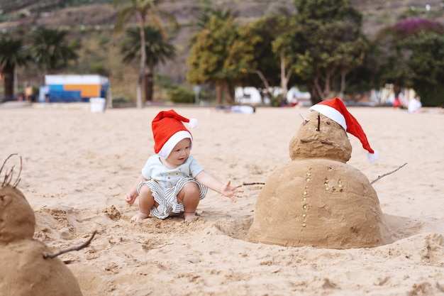 Kleines Mädchen, das mit sandigem Schneemann des neuen Jahres oder der frohen Weihnachten am tropischen Ozeansandstrand spielt