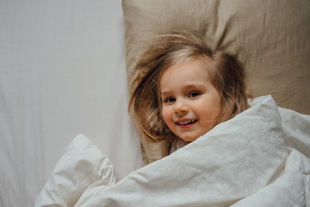 Kleines Mädchen, das morgens zu Hause mit einer Decke bedeckt im Bett liegt, wacht in ihrem Zimmer auf