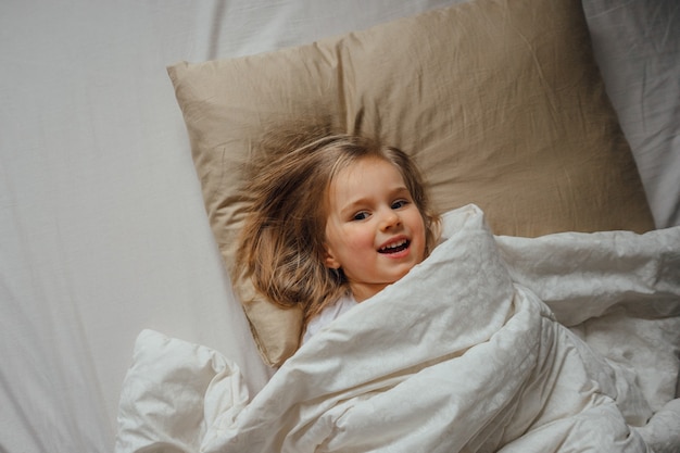 Kleines Mädchen, das morgens zu Hause mit einer Decke bedeckt im Bett liegt, wacht in ihrem Zimmer auf