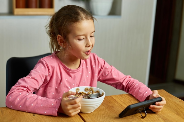 Kleines Mädchen, das Müsli mit Milch isst und Videos auf dem Smartphone ansieht