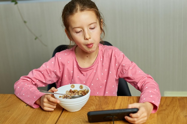 Kleines Mädchen, das Müsli mit Milch isst und Videos auf dem Smartphone ansieht