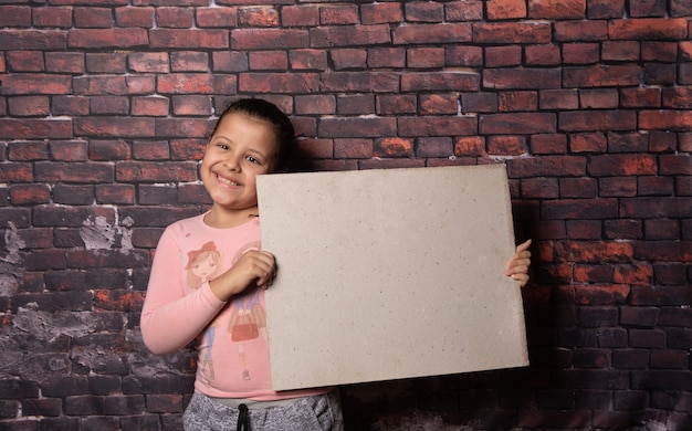 Kleines Mädchen, das Posen mit leeren Recyclingpapierbuchstaben vor einem alten Backsteinmauerhintergrund macht, dunkler Hintergrund, selektiver Fokus.