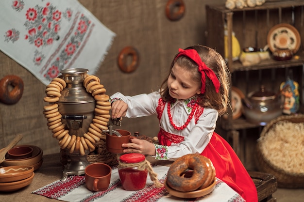 Kleines Mädchen, das rotes Stirnband und dekoratives Hemd trägt, das Tee von Samowar gießt, der Maslenitsa feiert