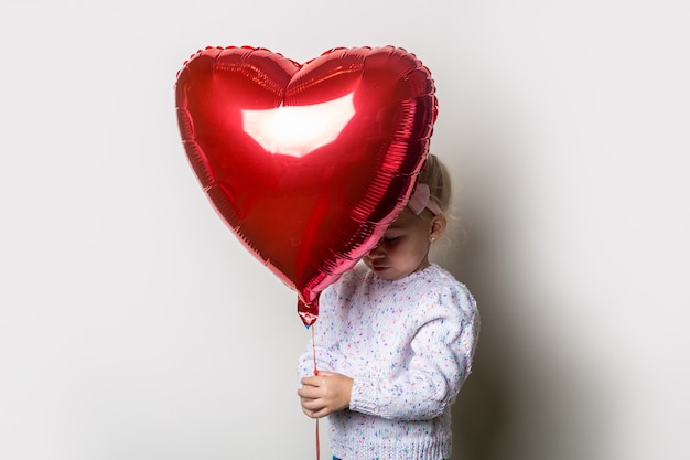 Kleines Mädchen, das sich hinter einem Herzluftballon auf einem hellen Hintergrund versteckt. Konzept für Valentinstag, Geburtstag. Banner.