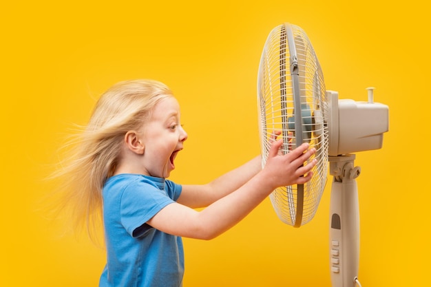 Kleines Mädchen, das sich im Wind bewegt, hat weiße Haare, hält einen Ventilator und schreit Kind und Ventilator auf gelbem Hintergrund
