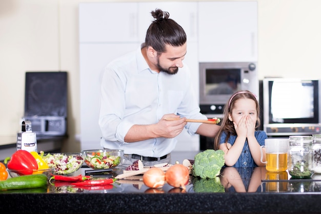 Kleines Mädchen, das sich weigert, Salat zu essen, während es zusammen mit Vater in der Küche kocht