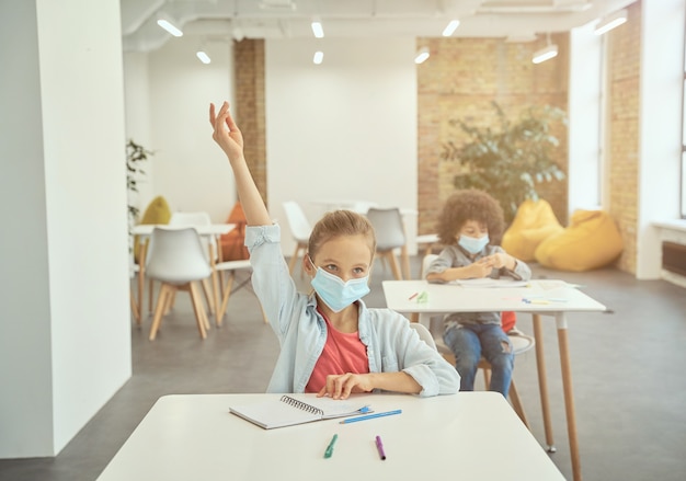 Kleines Mädchen, das während des Ausbruchs des Coronavirus eine schützende Gesichtsmaske trug, hob im Unterricht die Hand