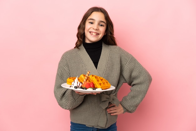 Kleines Mädchen, das Waffeln lokalisiert auf rosa Hintergrund hält, der mit Armen an der Hüfte aufwirft und lächelt