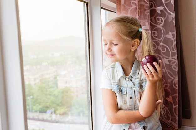 kleines Mädchen, das zu Hause auf dem Fensterbrett sitzt, im Fenster schaut und roten Apfel hält