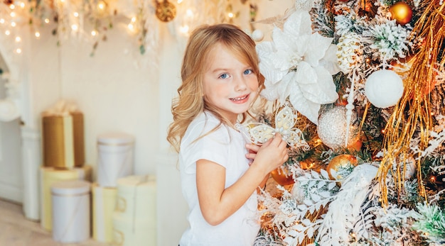 Kleines Mädchen, das zu Hause Weihnachtsbaum auf Weihnachtsabend verziert. Junge im hellen Raum mit Winterdekoration. Zeit für Feierkonzept Banner