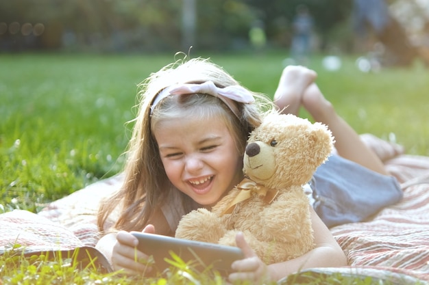 Kleines Mädchen, das zusammen mit ihrem Lieblings-Teddybär-Spielzeug draußen im Sommerpark in ihr Handy schaut.