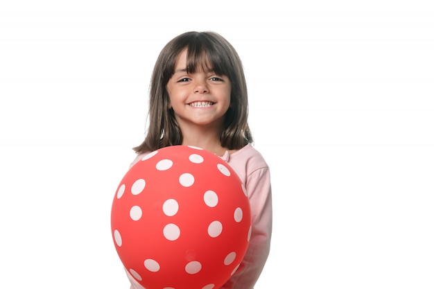 Kleines Mädchen des Brunette, das zur Kamera mit einem Ballon auf weißem Hintergrund lächelt