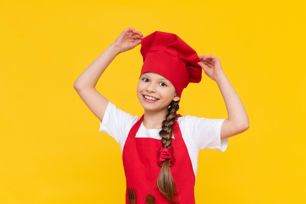 Kleines Mädchen des Küchenchefs wird Gerichte kochenDer Beruf eines Kochs für Kinder Kochen für Kinder Ein Kind in einer roten Schürze und einer Kappe auf einem gelb isolierten Hintergrund