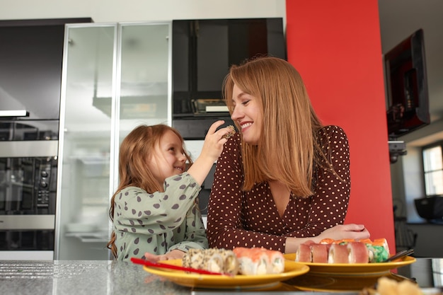 Kleines Mädchen füttert ihre Mutter Sushi mit Stäbchen aus nächster Nähe Essen zum Mitnehmen und Lieferservice Lachs-Sushi-Rollen