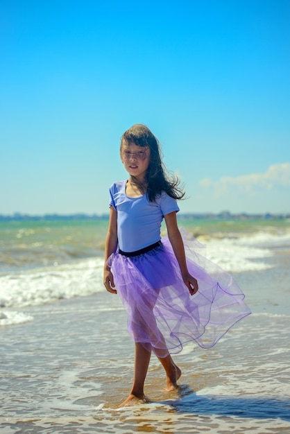 Kleines Mädchen geht an einem sonnigen Tag in einem lila Badeanzug auf dem Hintergrund der Wellen am Strand entlang