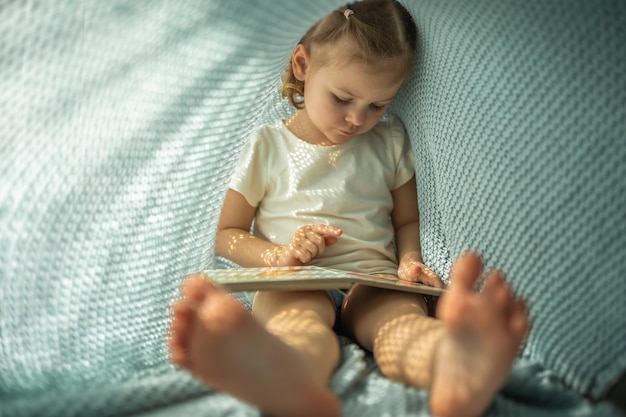 Kleines Mädchen genießt es, an einem sonnigen Morgen ein Buch unter blauem Strickkaro zu rezensieren