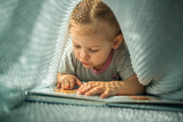 Kleines Mädchen genießt es, an einem sonnigen Morgen ein Buch unter blauem Strickkaro zu rezensieren