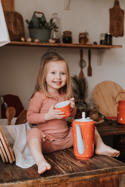 Kleines Mädchen gießt Tee in eine Tasse aus einer Teekanne aus rotem Keramikgeschirr in einer Holzküche mit weißen Erbsen