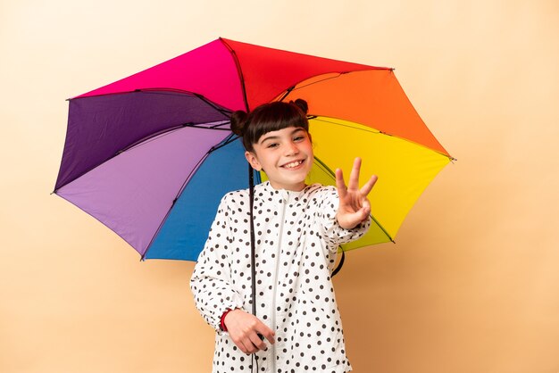 Kleines Mädchen hält einen Regenschirm lokalisiert auf beige glücklich und zählt drei mit den Fingern