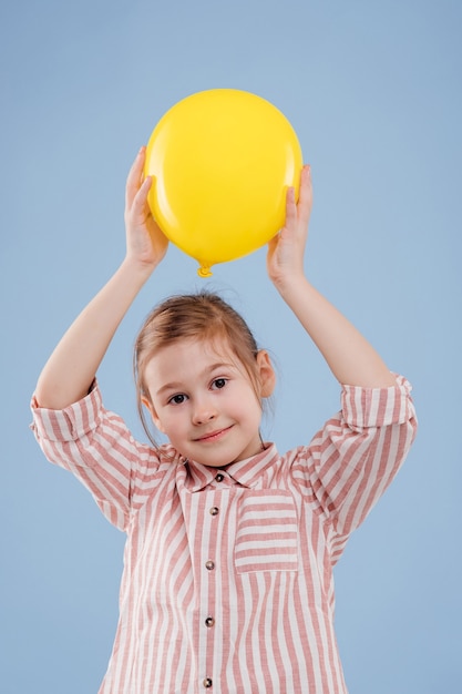 Kleines Mädchen hält gelben Ballon hoch und schaut in die Kamera, gekleidet in gestreiftes Hemd, isoliert auf blauem Ba...