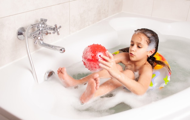 Kleines Mädchen hat Spaß in der Badewanne und spielt im aufblasbaren roten Ball, während es in der Badewanne sitzt