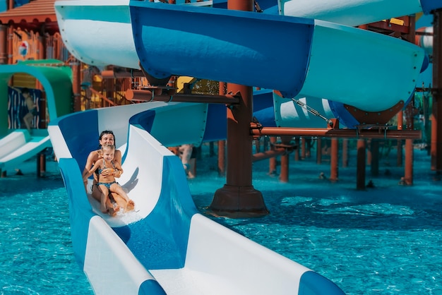 Kleines Mädchen im Badeanzug geht die blauen Rutschen hinunter zum Pool Mutter und Tochter spielen und schwimmen im Außenpool