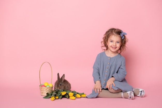 Kleines Mädchen im blauen Kleid mit Kaninchen, Tulpen, Korb mit bunten Eiern auf rosa Hintergrund.