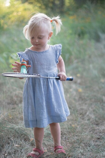 Kleines Mädchen im blauen Sommerkleid hält Badmintonschläger Dreijähriges Kind spielt Badminton Vertikaler Rahmen