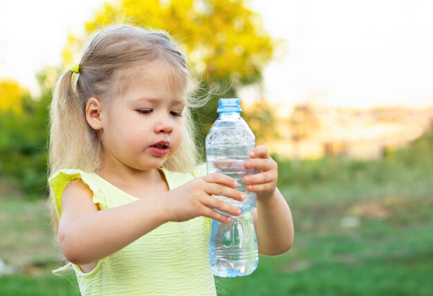 kleines Mädchen im gelben Kleid hält eine Flasche mit Trinkwasser auf Naturhintergrund