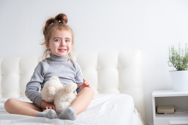 Kleines Mädchen im grauen Rollkragenpullover, der mit Teddybär auf weißer Bettwäsche spielt.
