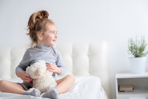 Kleines Mädchen im grauen Rollkragenpullover mit Teddybär, der lächelnd auf Bett sitzt.