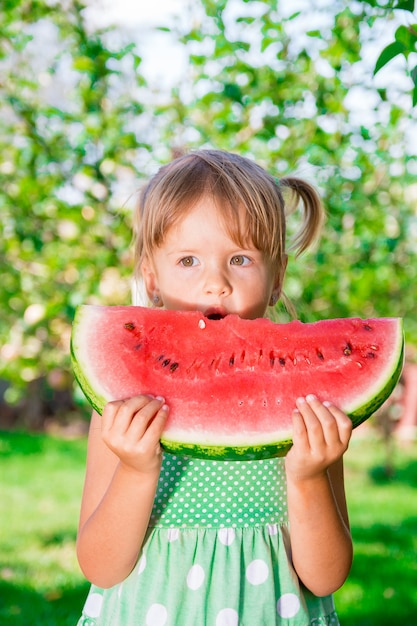 Kleines Mädchen im grünen Kleid, das große Scheibe Wassermelone im Park isst. Sommerzeit. Glücklich. Freude.