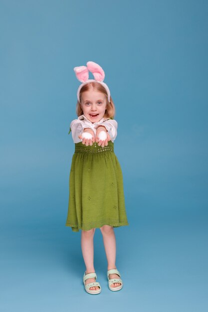 Kleines Mädchen im grünen Kleid und in den Hasenohren zeigt Huhn-Ostereier auf einer blauen Oberfläche