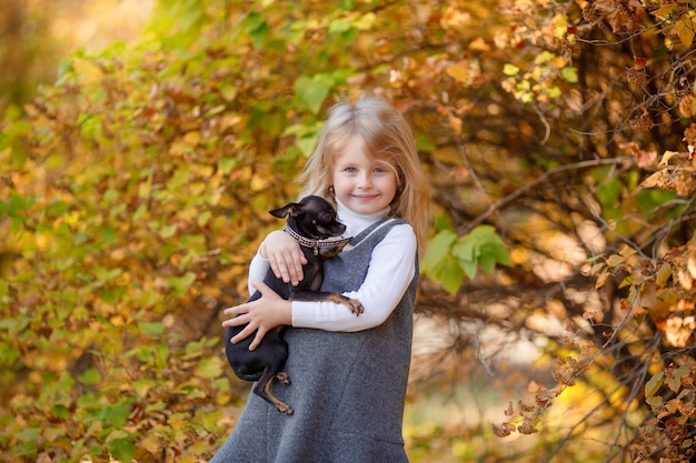 Kleines Mädchen im Herbst im Park, der einen kleinen Hund hält, lächelt