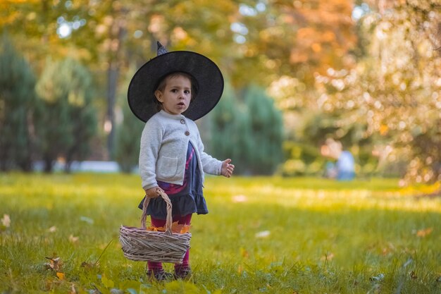 Kleines Mädchen im Hexenkostüm an Halloween im Herbstpark mit Korb voller gelber Blätter Kindheit