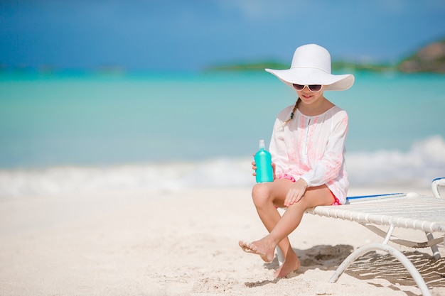 Kleines Mädchen im Hut mit der Flasche Sonnencreme sitzend am tropischen Strand