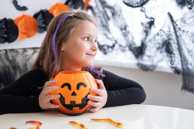 kleines Mädchen im Kostüm der Hexe, die Kürbisbuchse mit Süßigkeiten hält und Halloween zu Hause feiert