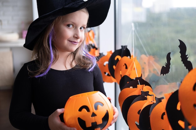 kleines Mädchen im Kostüm der Hexe, die Kürbisbuchse mit Süßigkeiten hält und Halloween zu Hause feiert