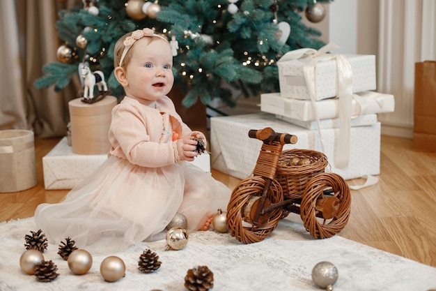 Kleines Mädchen im pfirsichfarbenen Kleid, das zu Hause neben dem Weihnachtsbaum sitzt