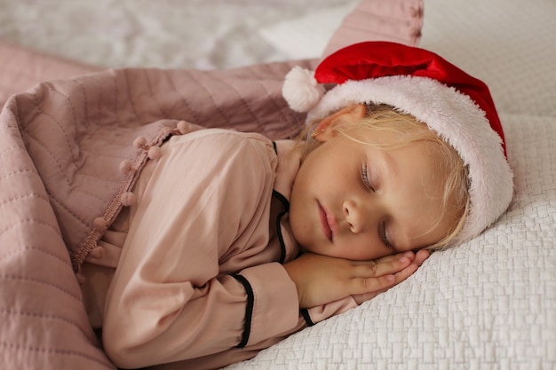 kleines Mädchen im rosa Pyjama und Weihnachtsmann-Hut schläft an Heiligabend im Bett