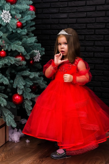 Kleines Mädchen im roten Kleid lächelt am Weihnachtsbaum. Kleines schönes Mädchen in einem roten Abendkleid